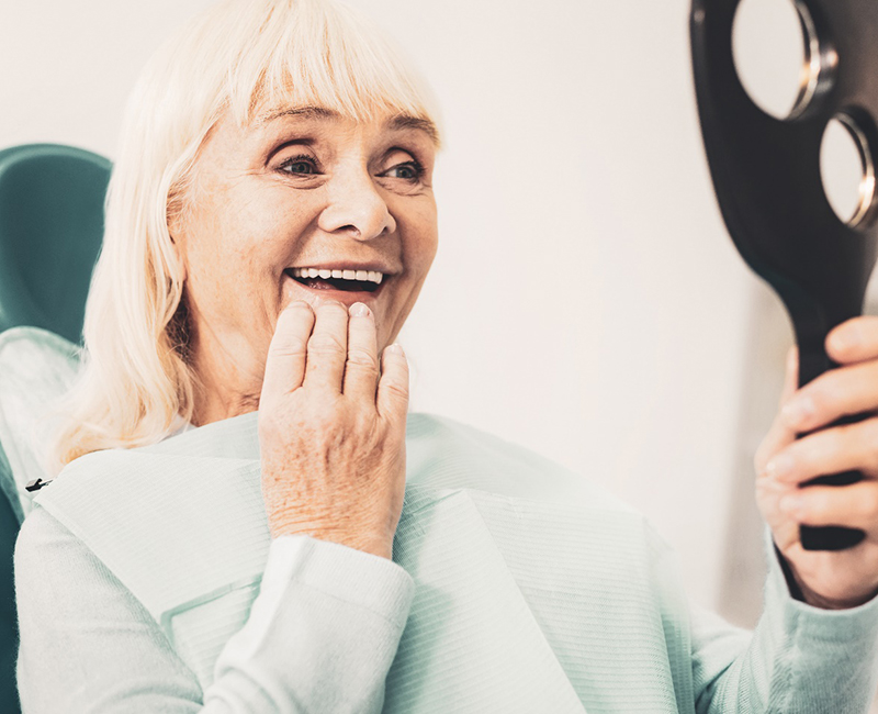 Senior woman looking in a handheld mirror with dentures in Rochester, MN